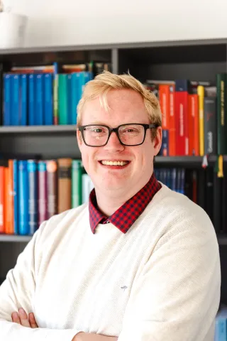 Das Bild zeigt Niklas Reinken, der vor einer Bücherwand mit bunten Büchern steht. Er hat blonde Haare und eine Brille. Er lächelt. Er trägt ein rot-kariertes Hemd und einen weißen Pullover.